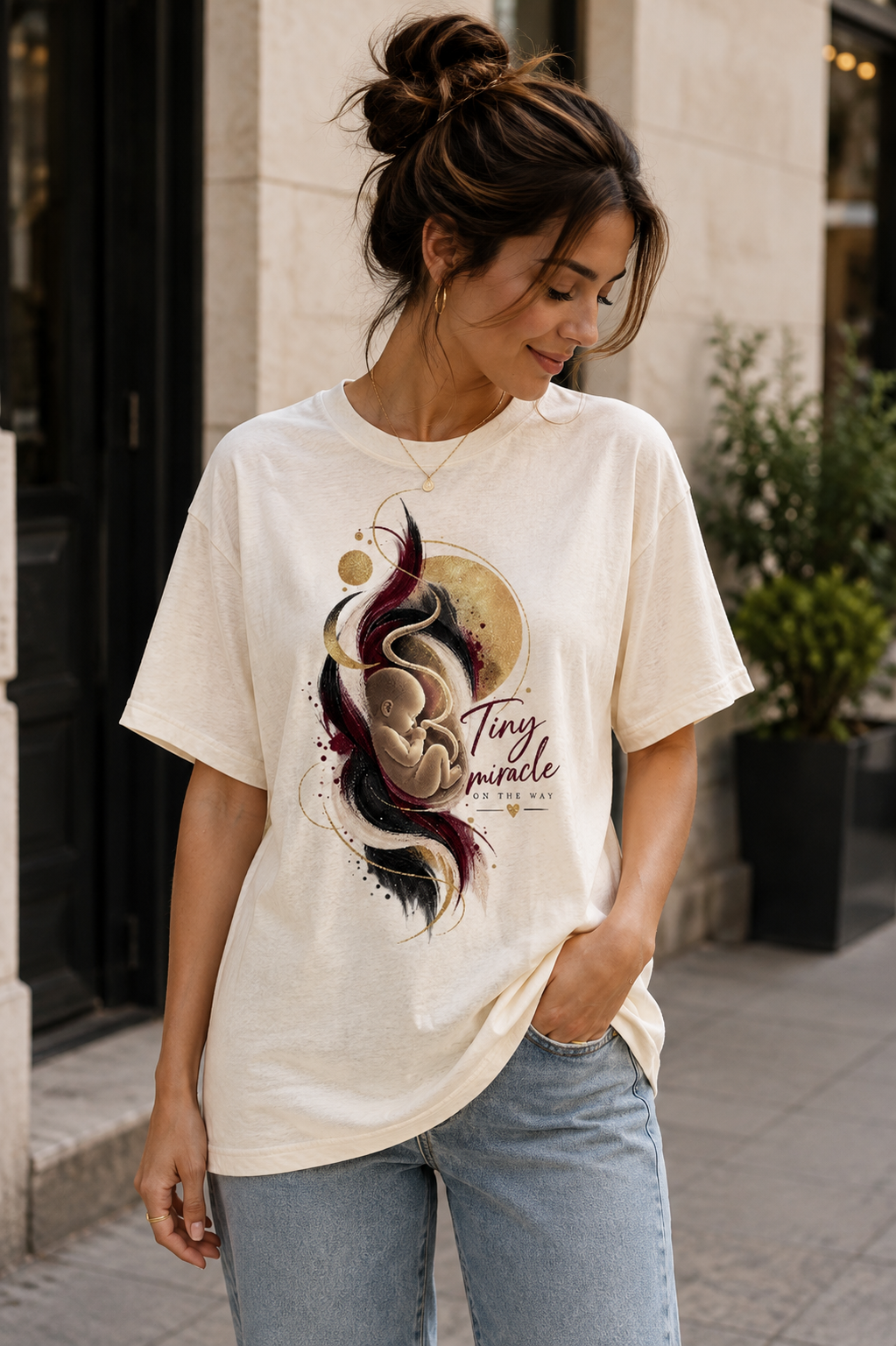 Cream women’s pregnancy announcement graphic T-shirt featuring an artistic baby illustration in burgundy, black and gold with the message Tiny miracle on the way.