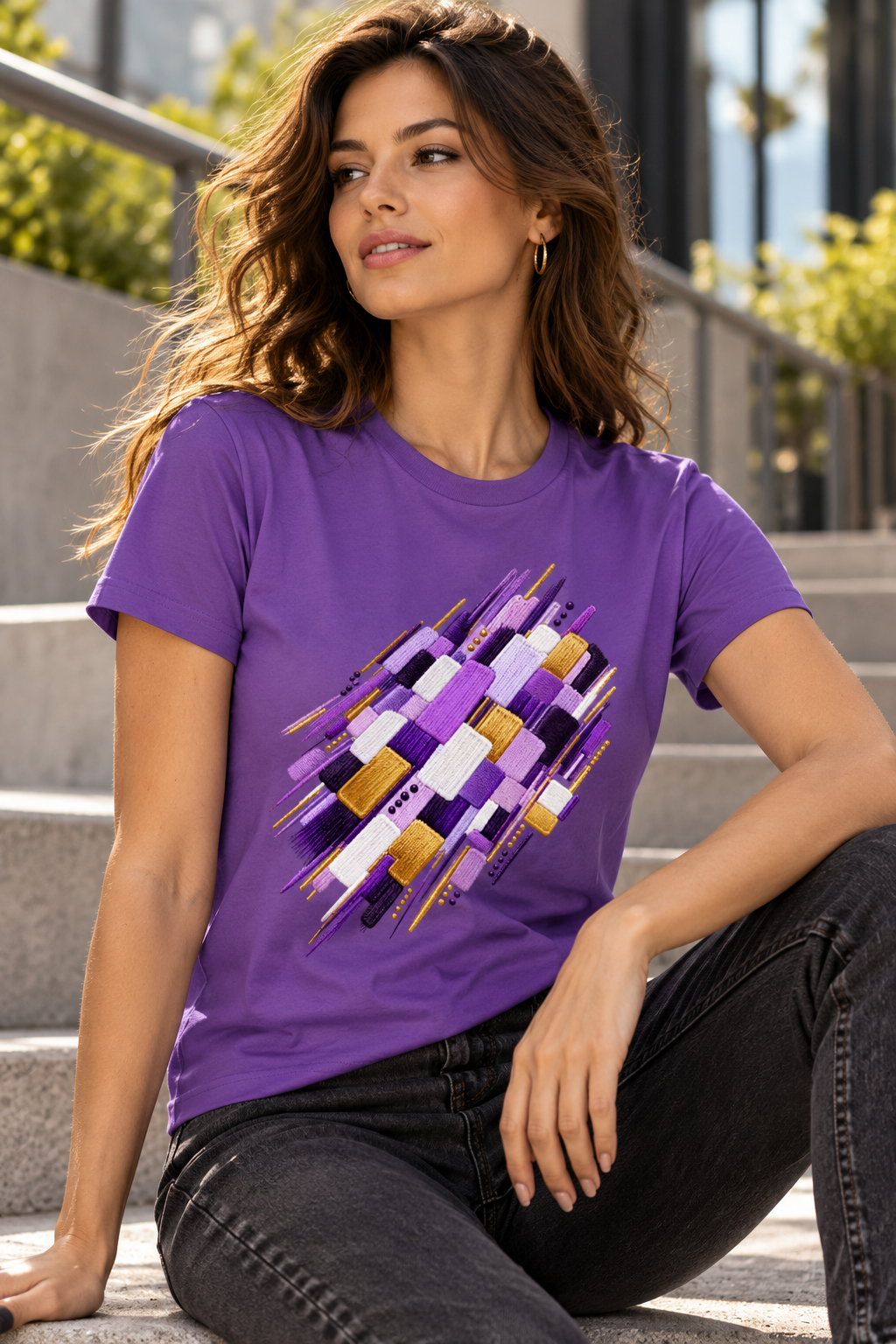 A woman sitting on outdoor concrete steps wears an Purple women's graphic T-shirt featuring abstract layered geometric artwork in violet, lilac, white, deep plum and gold by Exalyn Studio.